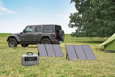 屋外でのキャンプ場で太陽パネルを広げてポータブル電源を充電している風景、背景に青空とジープ車